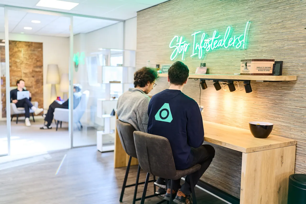 Due colleghi al bancone sotto l'insegna al neon “Stop Infostealers”